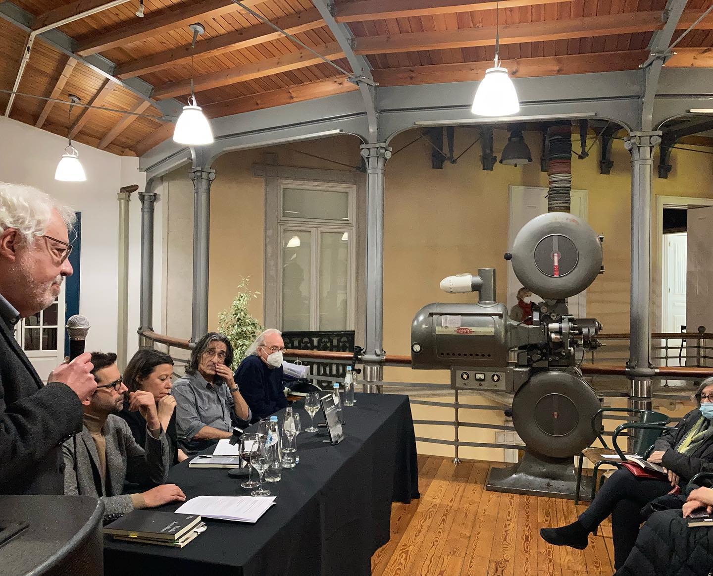 DIZER O MUNDO • CONVERSAS COM RUI NUNES E PAULO NOZOLINO • ALEXANDRA CARITA Sala com teto de madeira, mesa preta com pessoas, projetor de filme antigo e apresentação