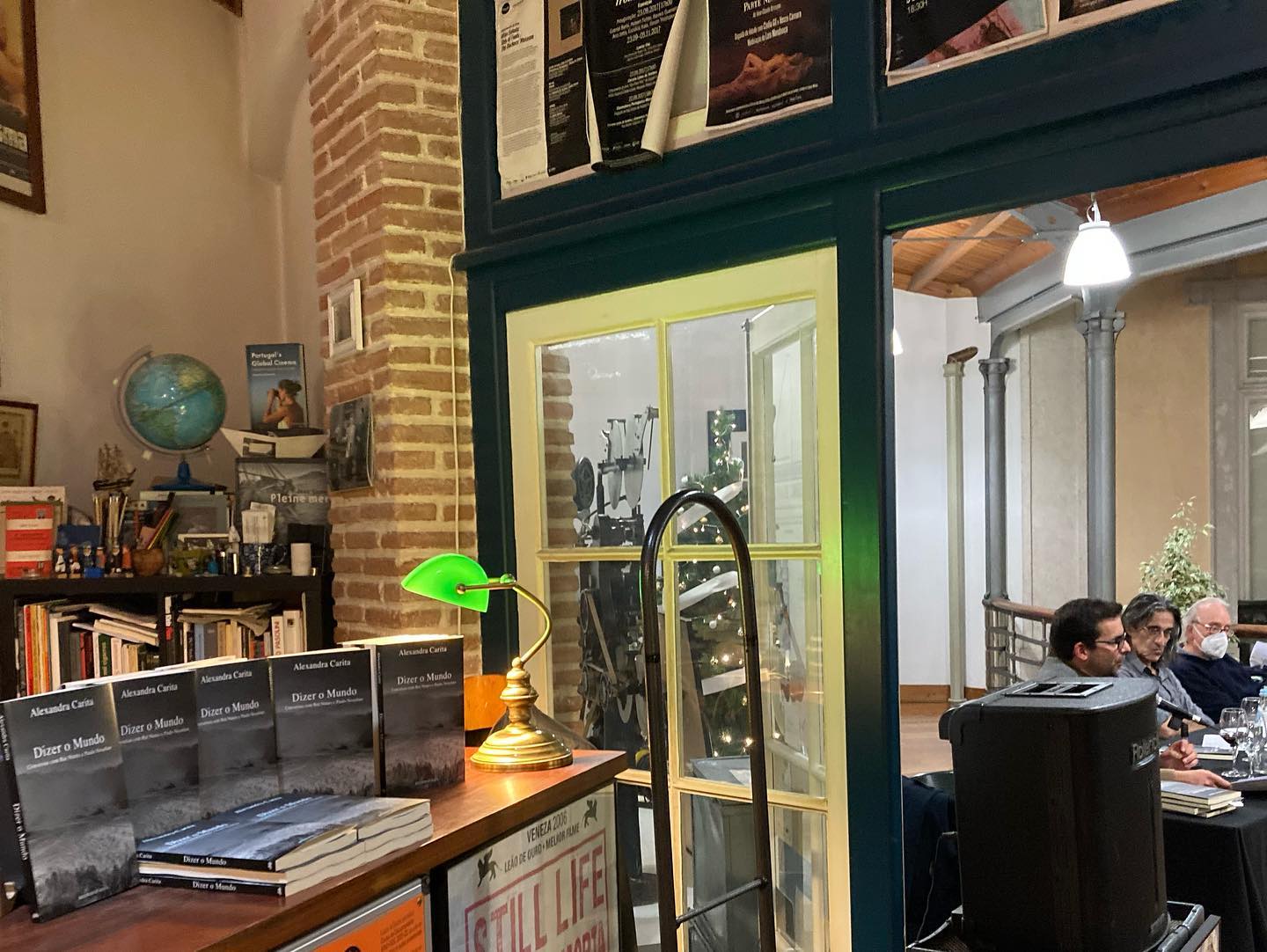 DIZER O MUNDO • CONVERSAS COM RUI NUNES E PAULO NOZOLINO • ALEXANDRA CARITA Mesa com vários exemplares do livro 'Diario Medina' em capa preta num espaço interior com elementos decorativos e pessoas ao fundo.