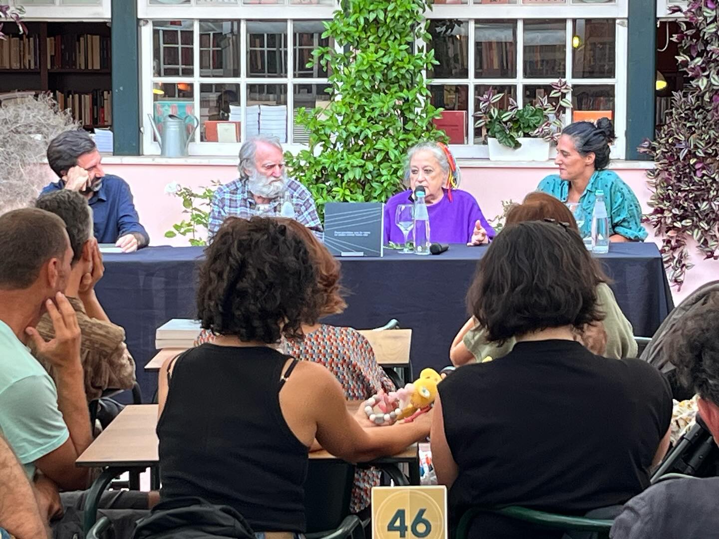 REGINA GUIMARÃES E SAGUENAIL ∙ DUAS PARALELAS QUE ÀS VEZES SE TOCAM OUTRAS VEZES NÃO ∙ INÊS SAPETA DIAS (ORG.) Evento ao ar livre com pessoas sentadas em volta de uma mesa preta e audiência