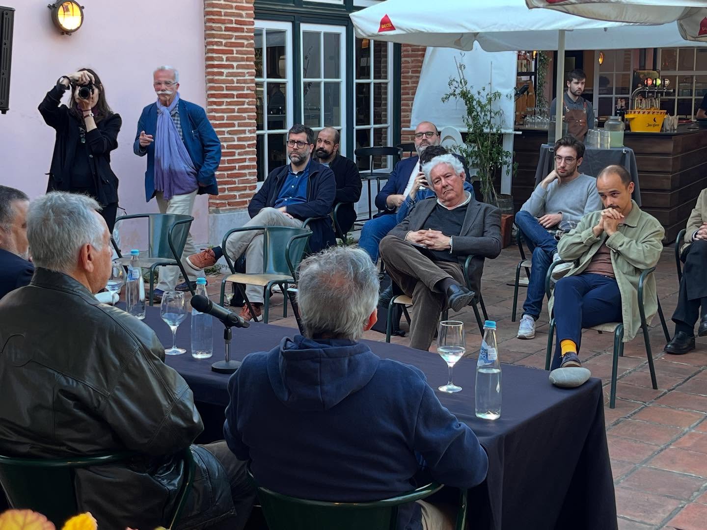 SOFIA E A EDUCAÇÃO SEXUAL ∙ EDUARDO GEADA Mesa preta com garrafas, copos e pessoas sentadas em cadeiras verdes num espaço exterior com piso de tijoleira.