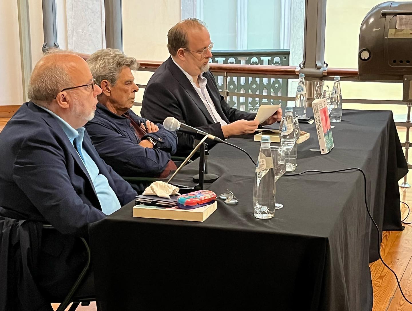 O CINEMA - UMA ESTÉTICA CRUA ∙ JOÃO PAULO DE OLIVEIRA CRUZ MENDES Três homens sentados atrás de mesa preta com copos e livro em cima