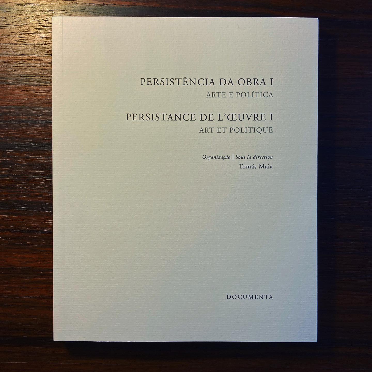 PERSISTÊNCIA DA OBRA I • ARTE E POLÍTICA • TOMÁS MAIA (ORG.) Livro branco com texto em português e francês na capa, sobre mesa de madeira