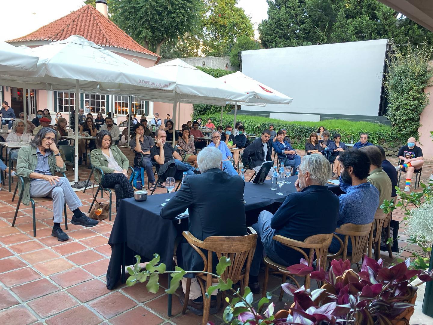 VITALINA VARELA • CADERNO DE RODAGEM • PEDRO COSTA Pessoas sentadas em espaço exterior com guarda-sóis e mesa preta para reunião