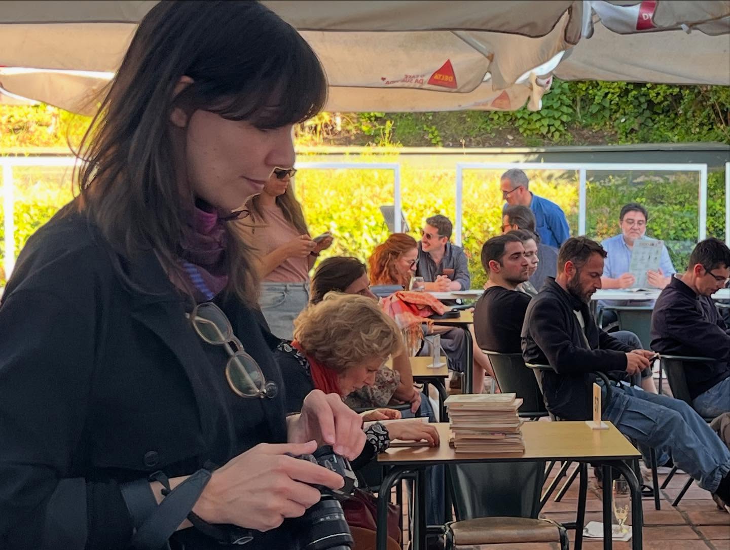 SOFIA E A EDUCAÇÃO SEXUAL ∙ EDUARDO GEADA Pessoa com casaco preto segura câmara numa esplanada com várias pessoas sentadas