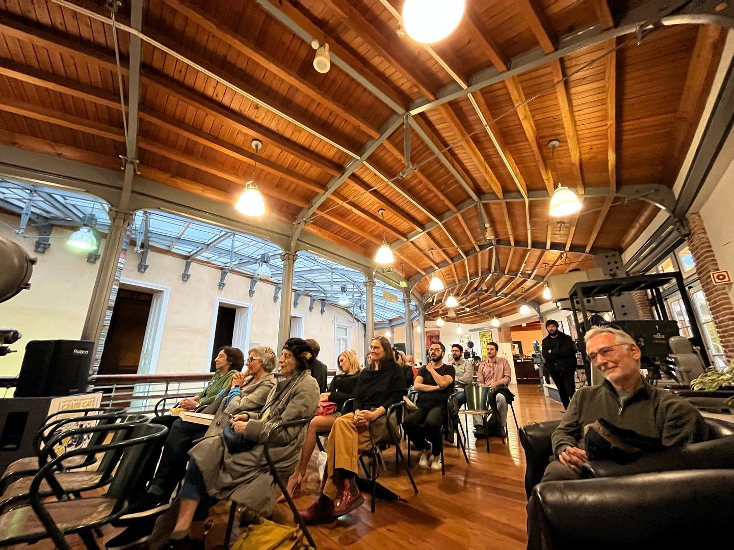 O CINEMA - UMA ESTÉTICA CRUA ∙ JOÃO PAULO DE OLIVEIRA CRUZ MENDES Pessoas sentadas em cadeiras numa sala interior com teto de madeira curvado e luzes pendentes