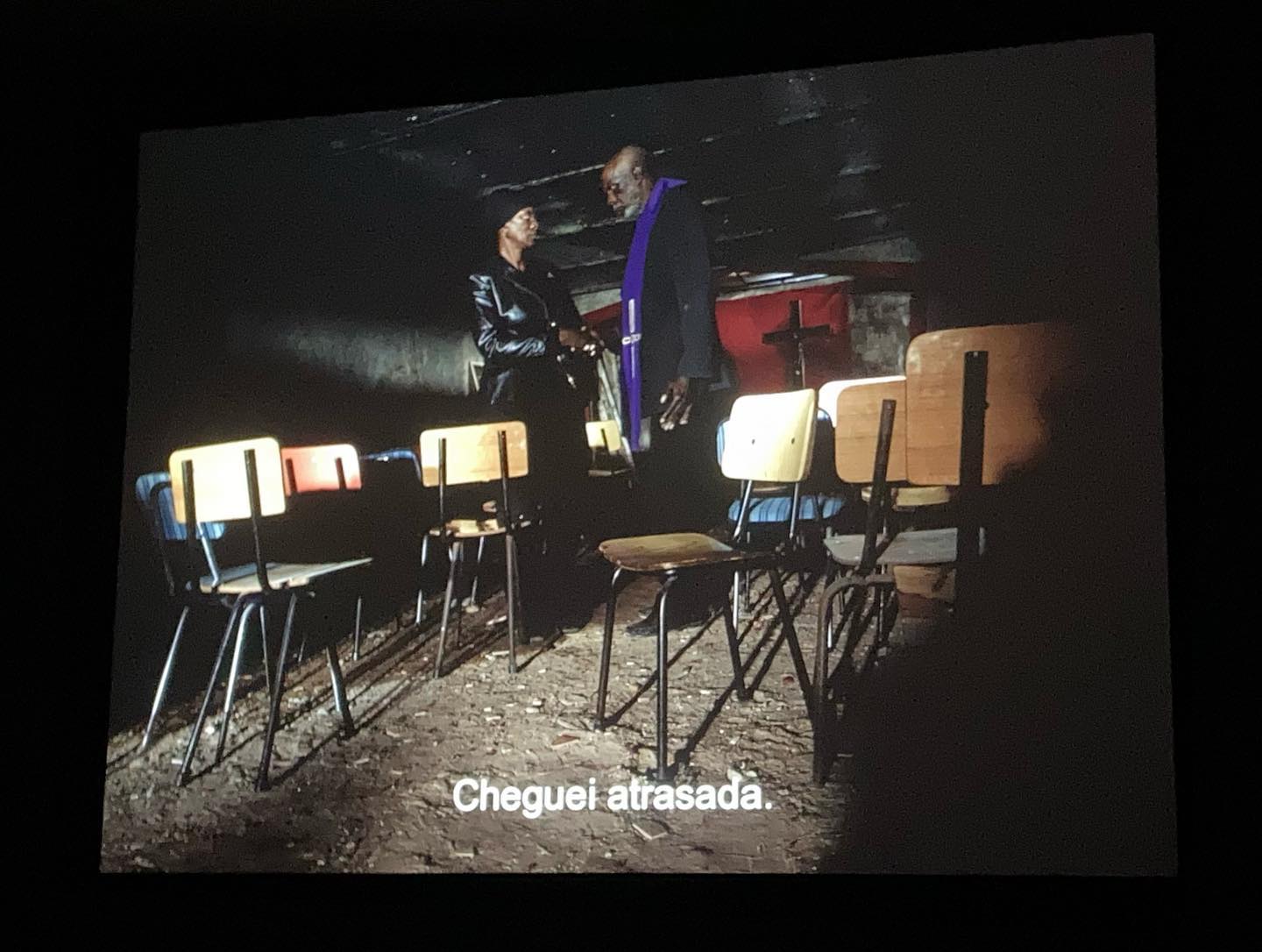 VITALINA VARELA • CADERNO DE RODAGEM • PEDRO COSTA Sala escura com cadeiras escolares e duas pessoas de pé ao fundo com legenda em português