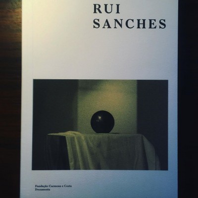 Capa de livro branco com texto RUI SANCHES e imagem de esfera preta sobre pano branco.