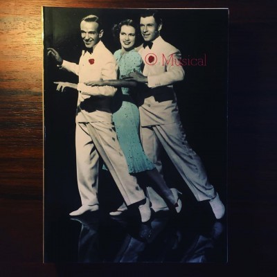 Capa de livro com três pessoas a dançar e o título 'Musical', fundo escuro, superfície em madeira