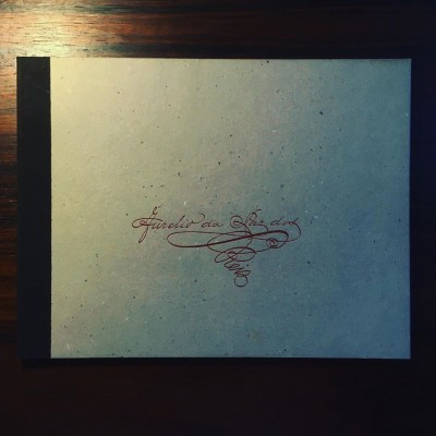 Caderno bege com lombada preta e texto manuscrito vermelho no centro da capa