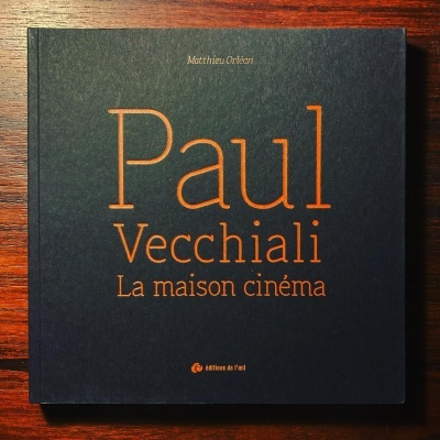 Livro preto com texto dourado 'Paul Vecchiali La maison cinéma' sobre mesa de madeira