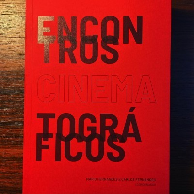 Livro vermelho com texto 'ENCONTROS CINEMATOGRÁFICOS' em preto e cinza sobre uma mesa de madeira