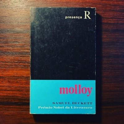 Livro 'Molloy' de Samuel Beckett com capa preta e faixa azul sobre madeira escura
