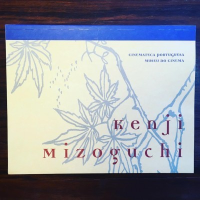 Envelope amarelo claro com folhas cinzentas e texto em azul e vermelho sobre mesa de madeira.