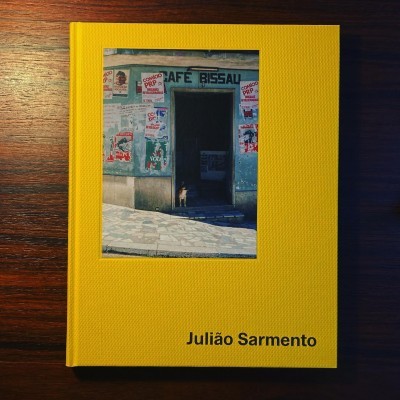 Capa de livro amarela com fotografia de porta e cartazes e nome Julião Sarmento