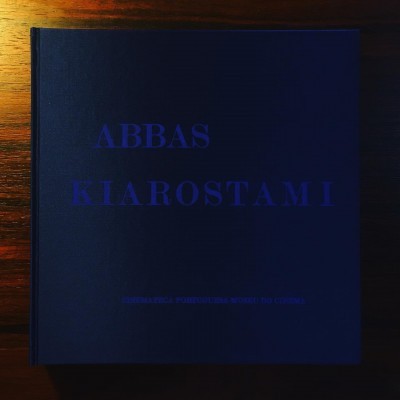 Livro preto com texto azul ABBAS KIAROSTAMI na capa sobre madeira