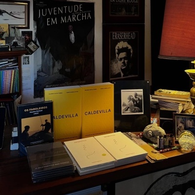 Mesa com livros, fotografias, DVDs e candeeiro em cima, com posters de filmes na parede