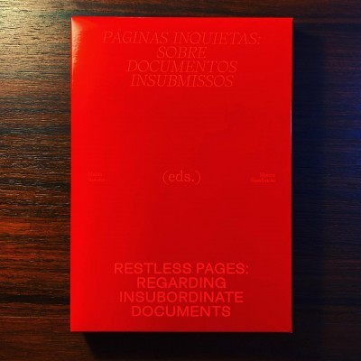 Livro vermelho com texto em relevo em português e inglês sobre documentos insubmissos numa mesa de madeira escura