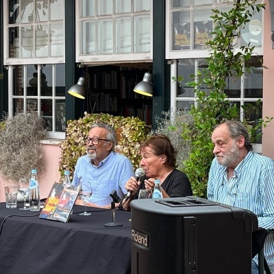 Mesa preta com microfones e três pessoas sentadas em evento exterior junto a janelas e plantas