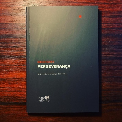 Capa de livro preta com texto branco e laranja sobre mesa de madeira escura