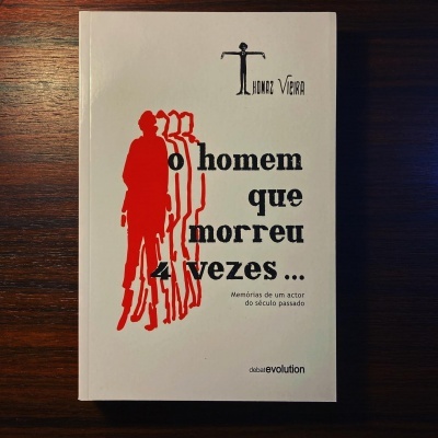 Capa de livro 'o homem que morreu 4 vezes...' com texto e silhuetas vermelhas sobre fundo branco e madeira.