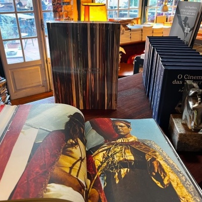 Livros expostos numa loja, um aberto com imagens de roupas históricas, outro com lombada padrão madeira, pilha de livros 'O Cinema das Mulheres' e escultura de rato