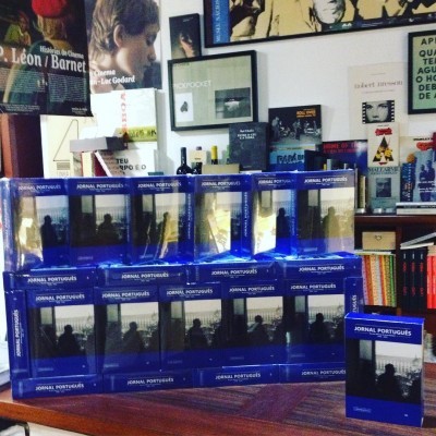 Caixas azuis 'JORNAL PORTUGUÊS' empilhadas numa livraria com livros e cartazes na parede