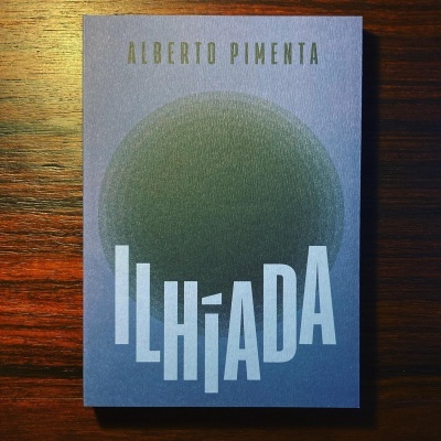 Livro ILHÍADA azul com círculo verde escuro e texto em branco e dourado sobre mesa de madeira