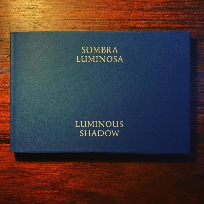 Caixa azul escura com texto dourado Sombra Luminosa Luminous Shadow
