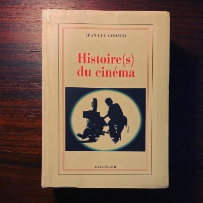 Livro 'Histoire(s) du cinéma' de Jean-Luc Godard, capa bege com ilustração em silhueta de câmera e operador