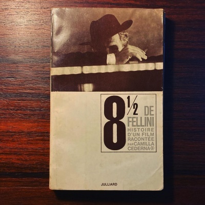 Livro '8 1/2 de Fellini' com capa branca e castanha, fotografias e texto impressos sobre madeira escura