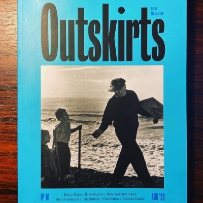 Capa da revista Outskirts com fotografia a preto e branco de duas pessoas na praia e fundo azul