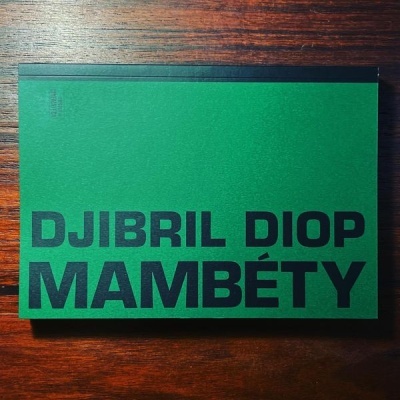 Livro com capa verde e texto preto DJIBRIL DIOP MAMBÉTY sobre madeira escura