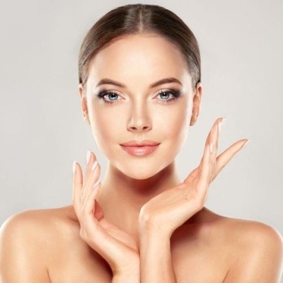 Rosto de mulher jovem com pele clara e cabelo castanho, maquilhagem natural, mãos próximas do rosto em fundo cinza claro