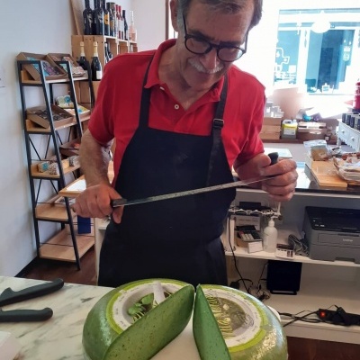 Roda de queijo verde cortada ao meio numa bancada branca e homem a medir o queijo com régua.