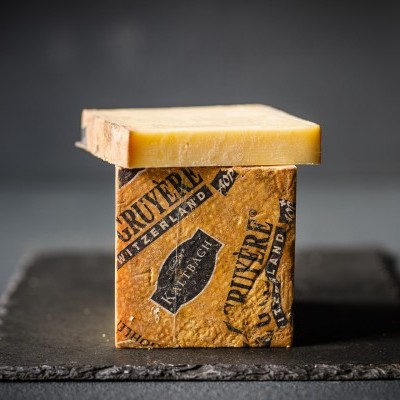 Queijo suíço Gruyere parcialmente embalado em papel castanho com texto preto, sobre superfície escura