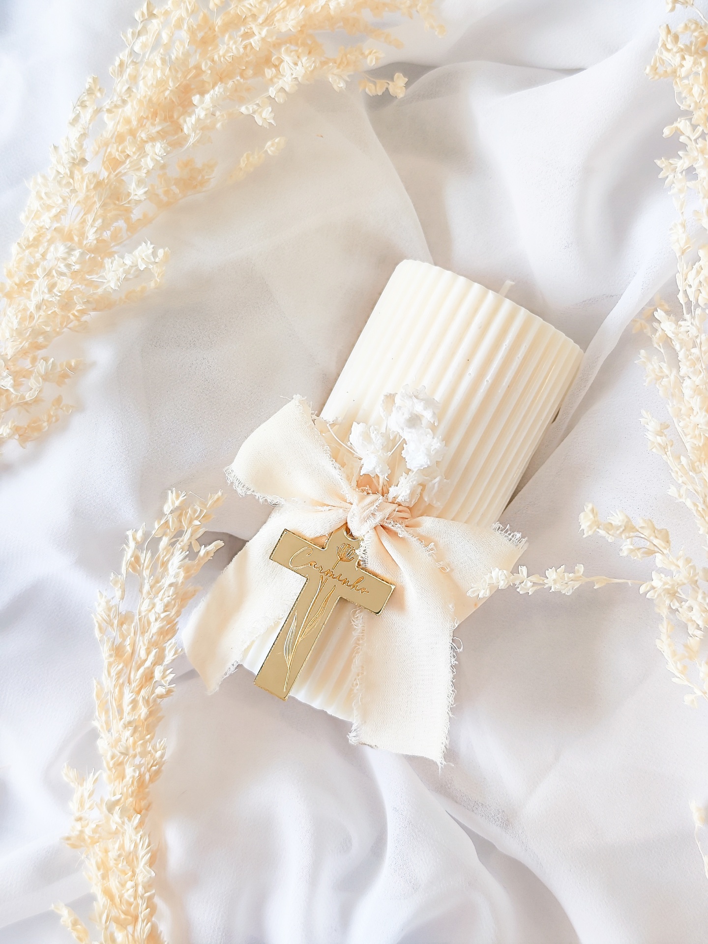 Vela branca canelada com decoração de laço, cruz dourada e flores secas sobre tecido branco.