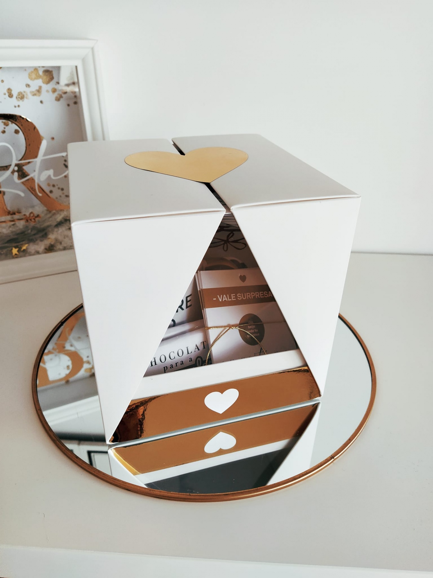 Caixa branca com coração dourado, sobre espelho redondo dourado, com vale-surpresa e embalagem de chocolate visíveis.