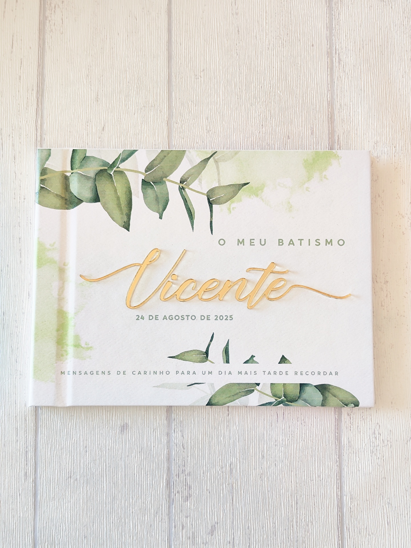 Livro de batismo com capa branca, folhas verdes e texto dourado e preto
