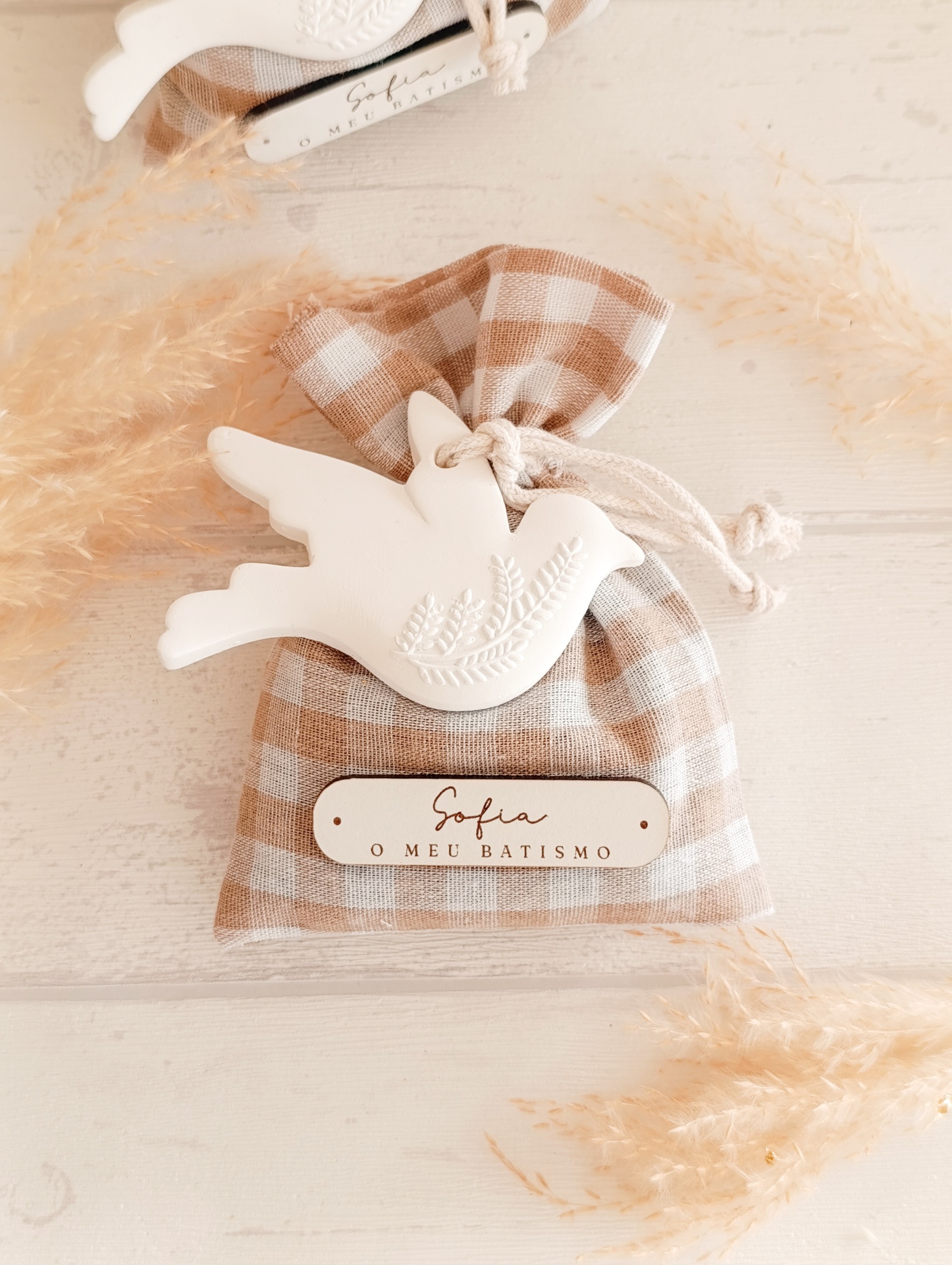 Saquinho de tecido xadrez castanho e branco com figura de pássaro branco e placa com texto de batismo