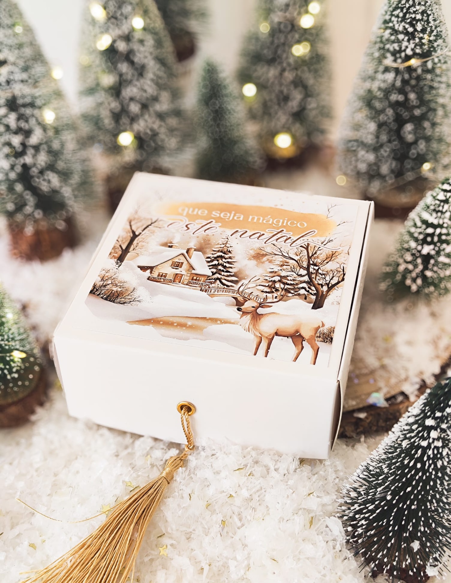 Caixa branca decorativa com ilustração natalícia e pompom dourado em ambiente com árvores de Natal