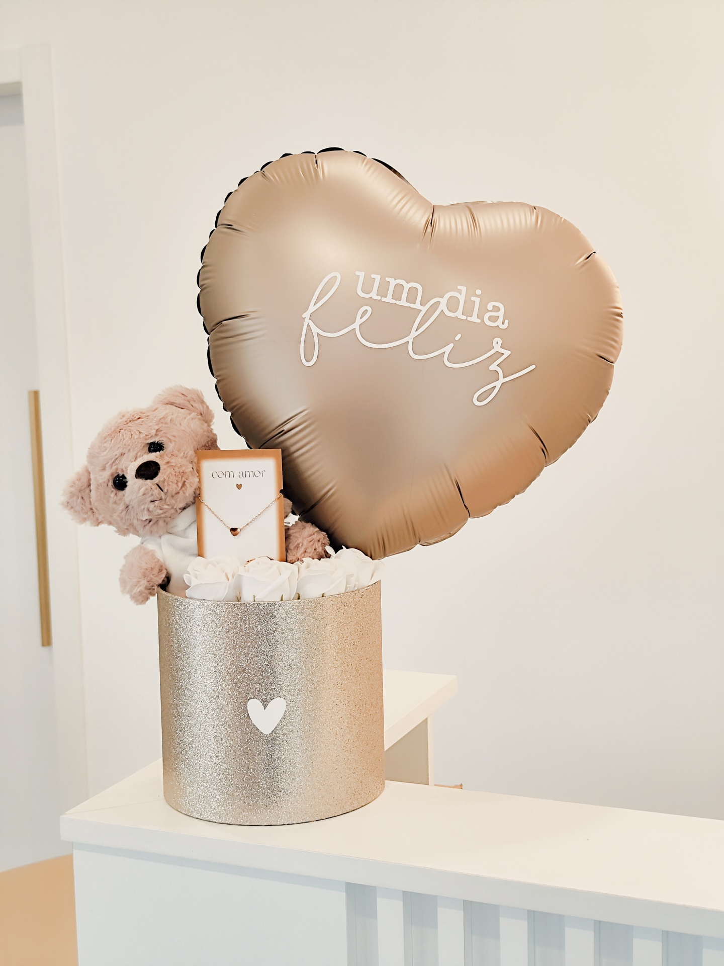 Conjunto de presente com balão dourado em forma de coração, ursinho, colar e flores brancas numa caixa dourada.