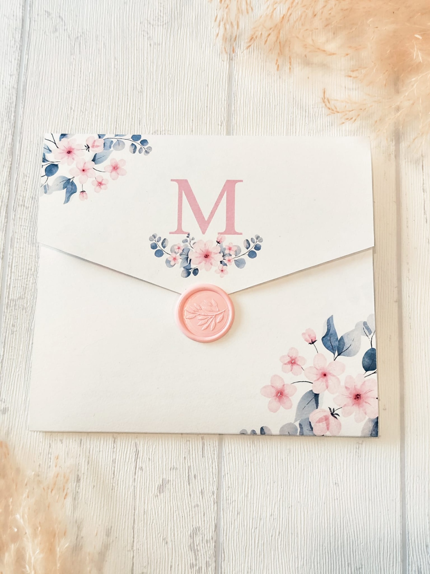 Envelope branco com flores rosas e azuis, selo de cera rosa e letra M