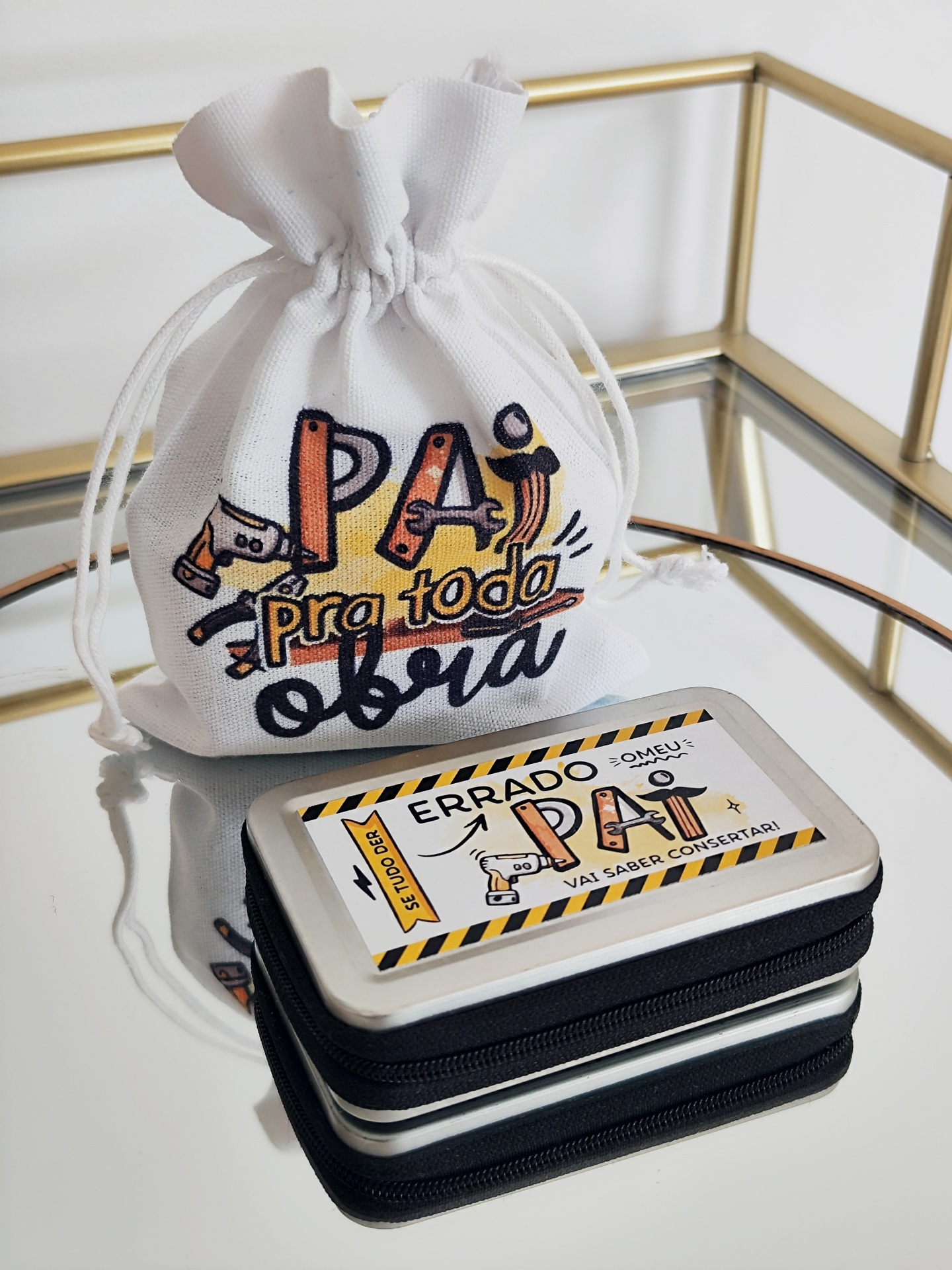 Saco branco estampado com mensagem para pai e caixa metalizada com texto humorístico, sobre superfície espelhada