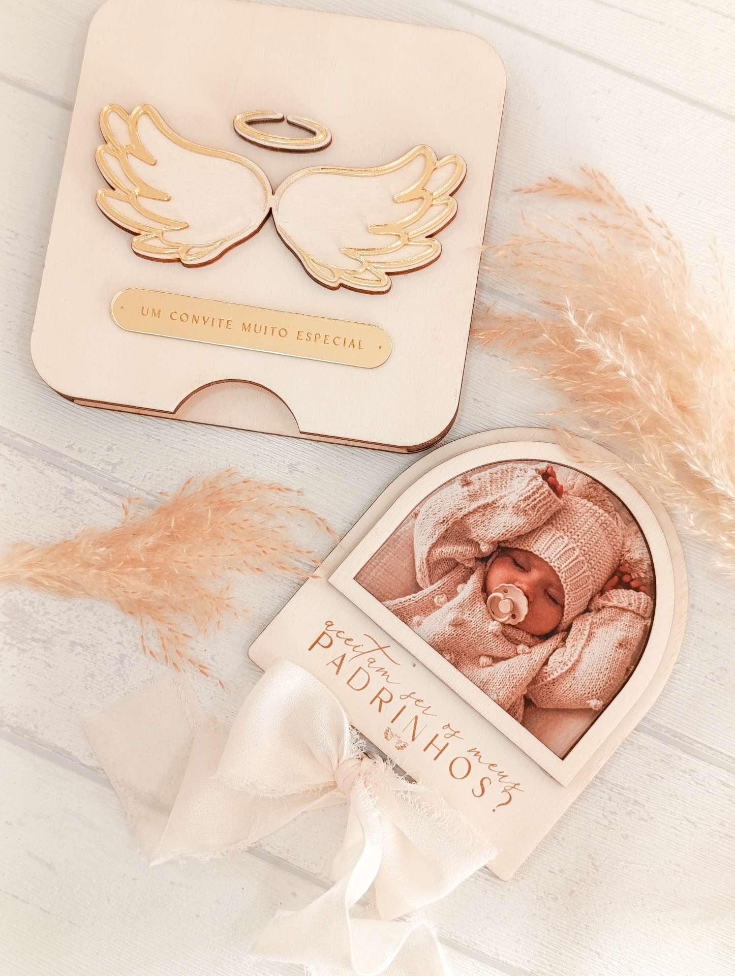 Convite de madeira claro com asas douradas e foto de bebé com laço branco