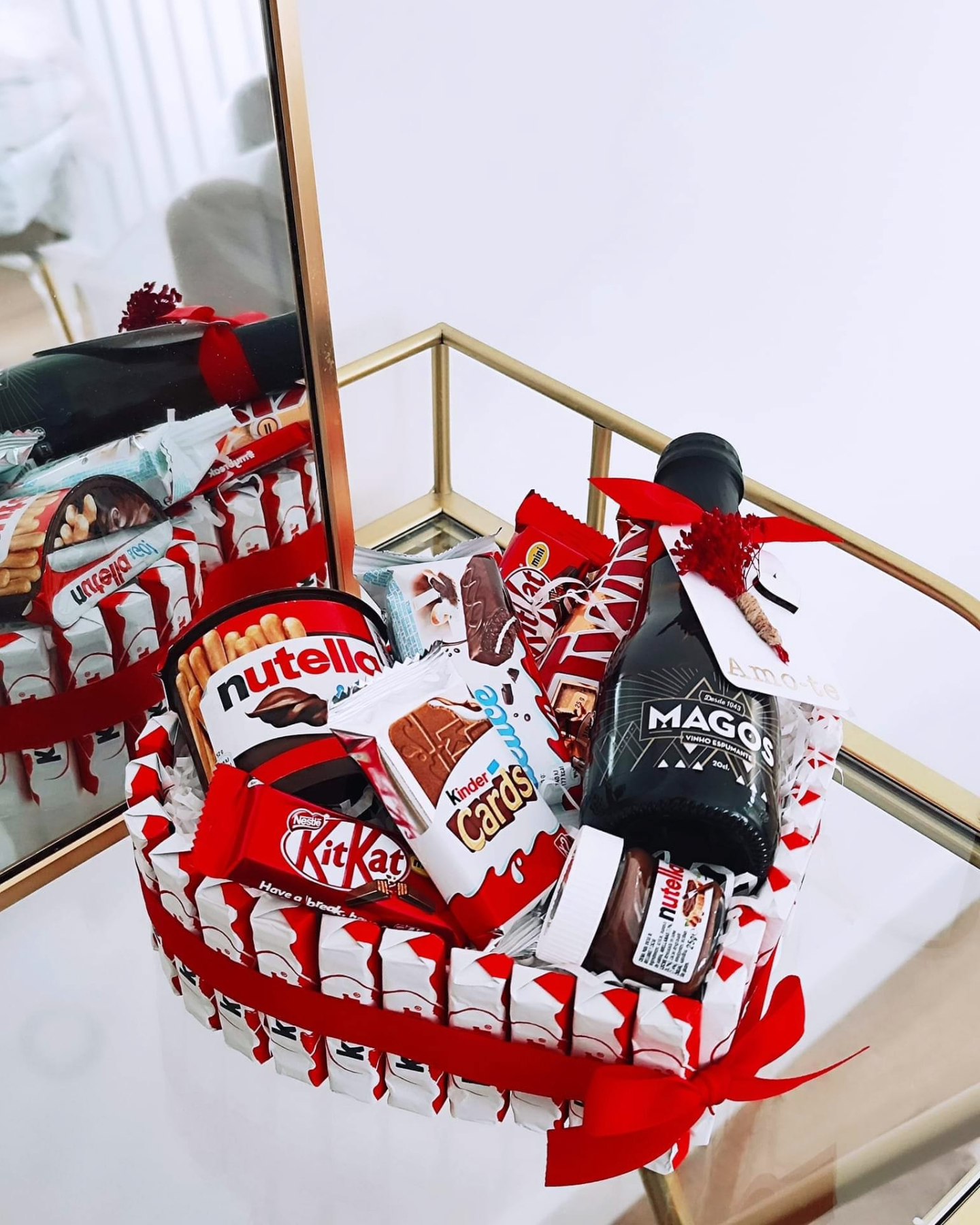 Cesta de presente com chocolates Kinder, Nutella, KitKat e garrafa de licor MAGOS sobre mesa espelhada