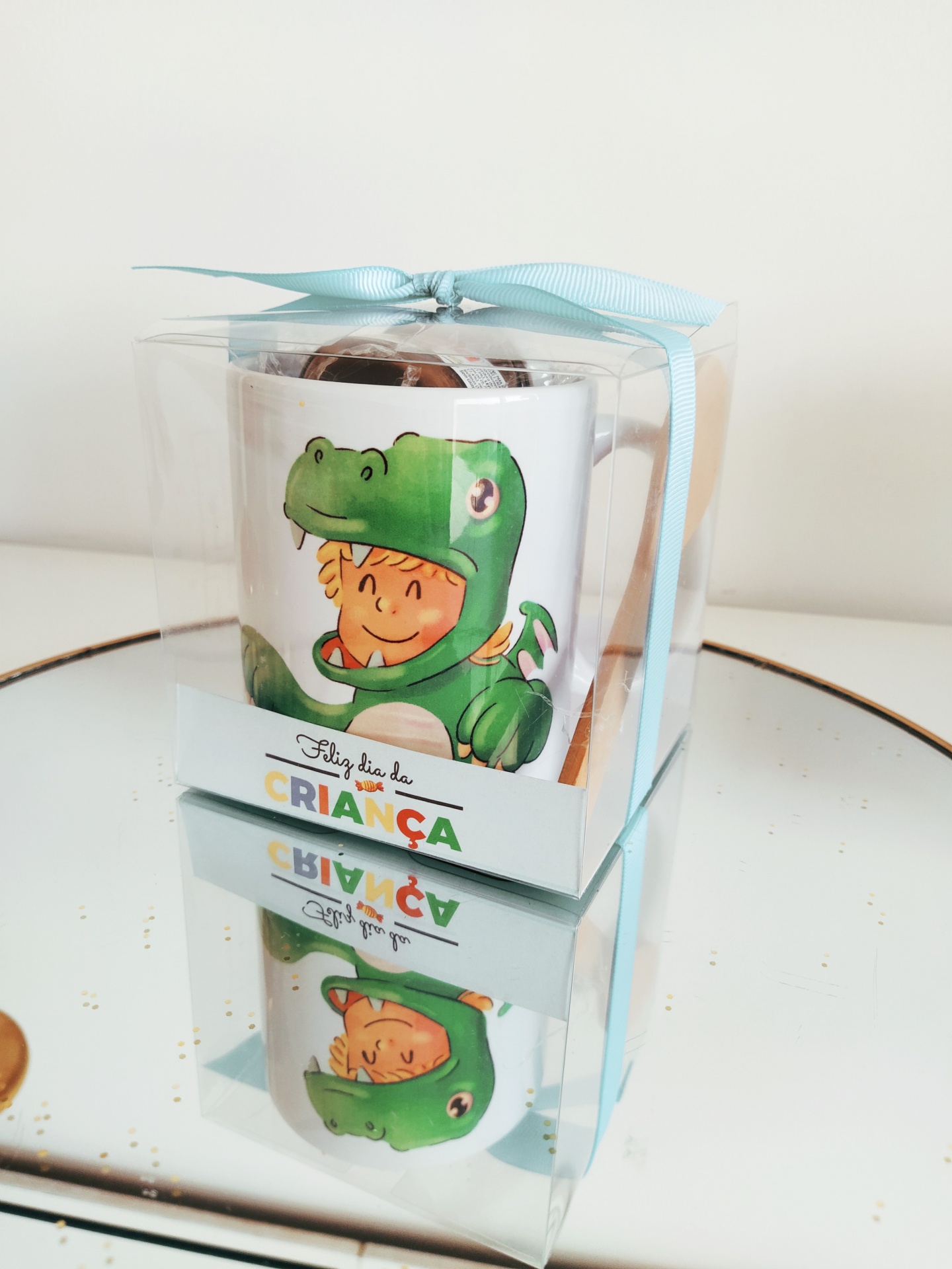 Caneca com ilustração de criança em fato de dinossauro embalada em caixa transparente com fita azul