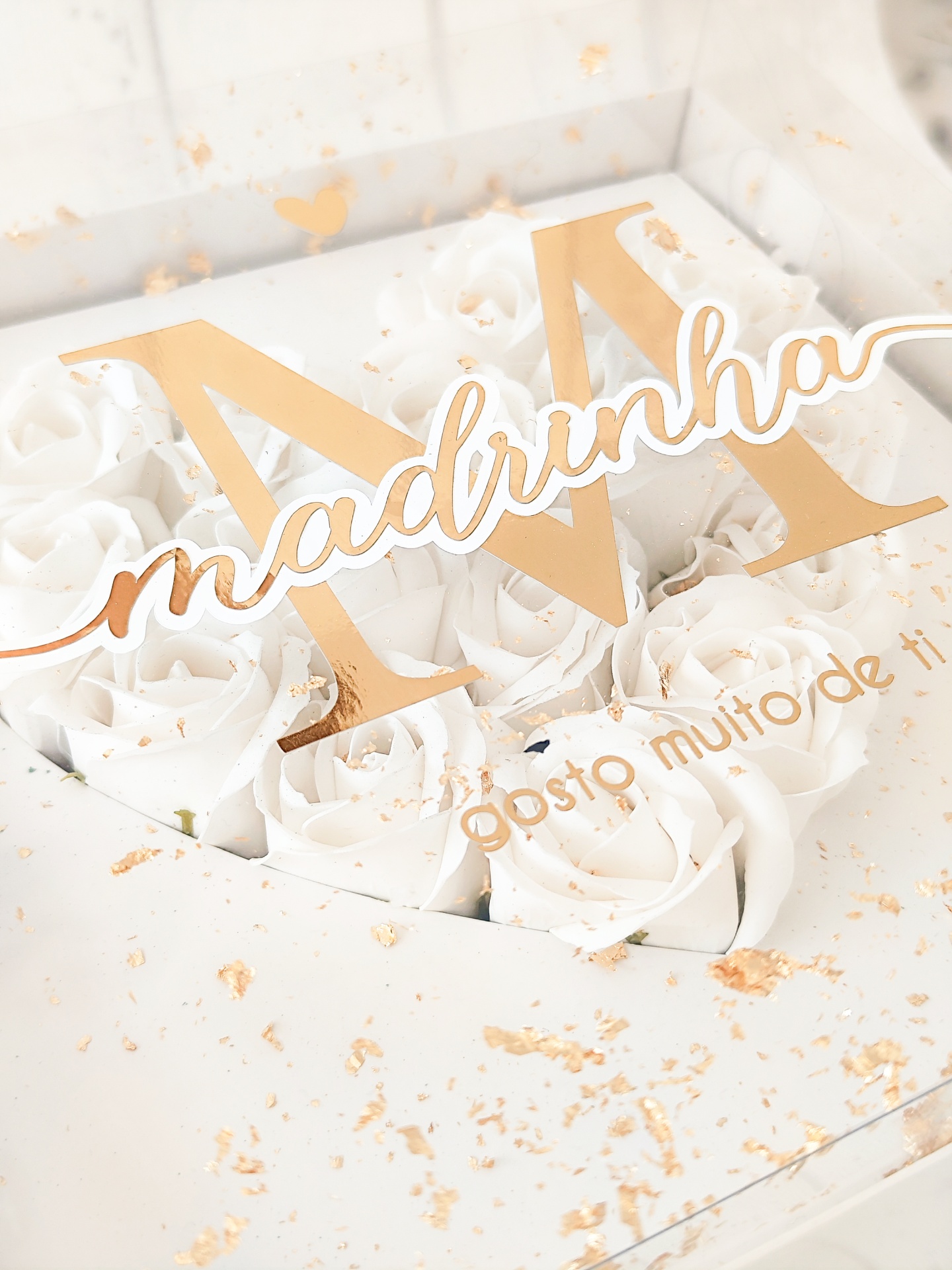 Caixa transparente com flores brancas artificiais, letra 'M' e texto dourado 'madrinha' e 'gosto muito de ti'