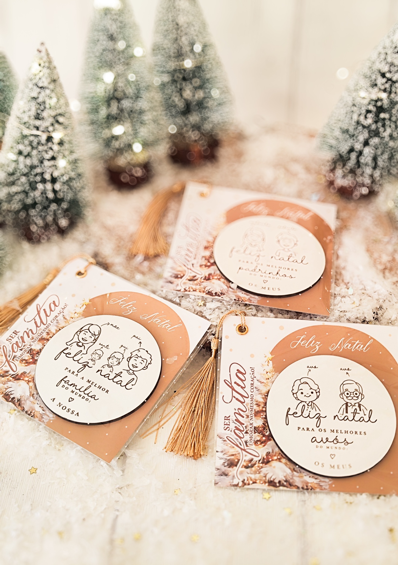 Enfeites de Natal brancos com ilustrações de família e texto em cartões castanhos com franjas douradas sobre neve artificial com pinheiros