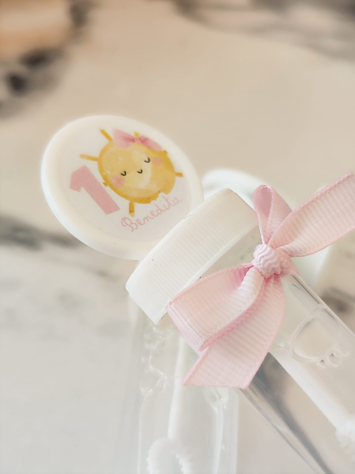 Garrafa transparente com tampa branca, laço rosa e etiqueta decorativa com sol e número 1