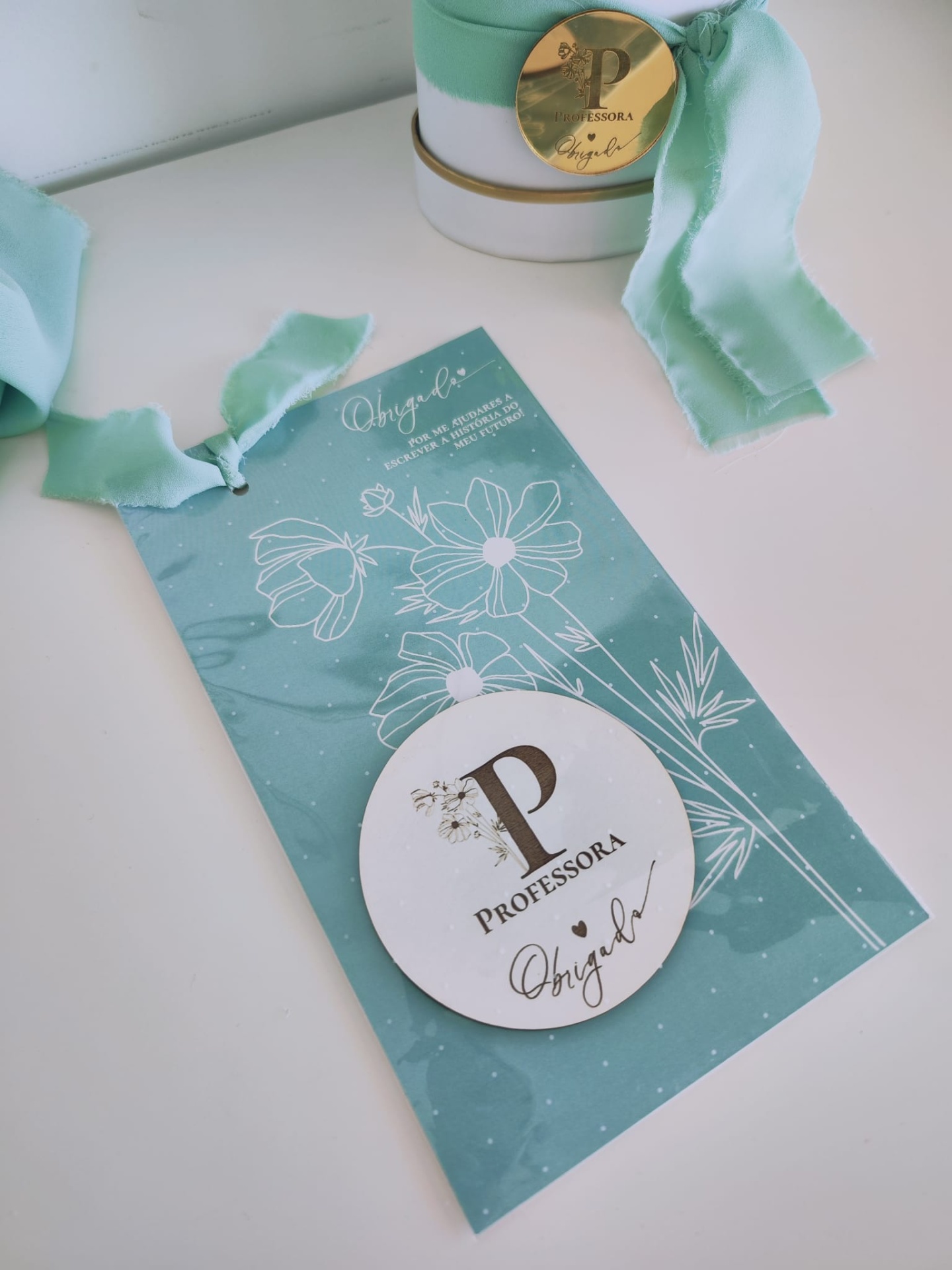 Etiqueta azul com medalha branca e texto para professora com recipiente azul e branco ao fundo